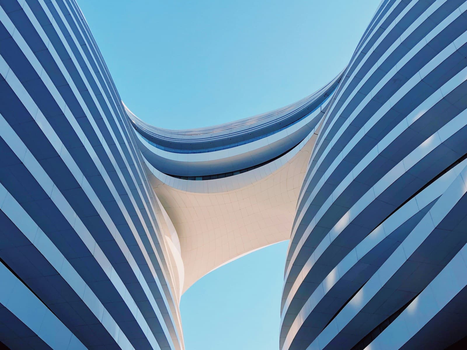 Looking up between two curved glass buildings toward a bright sky